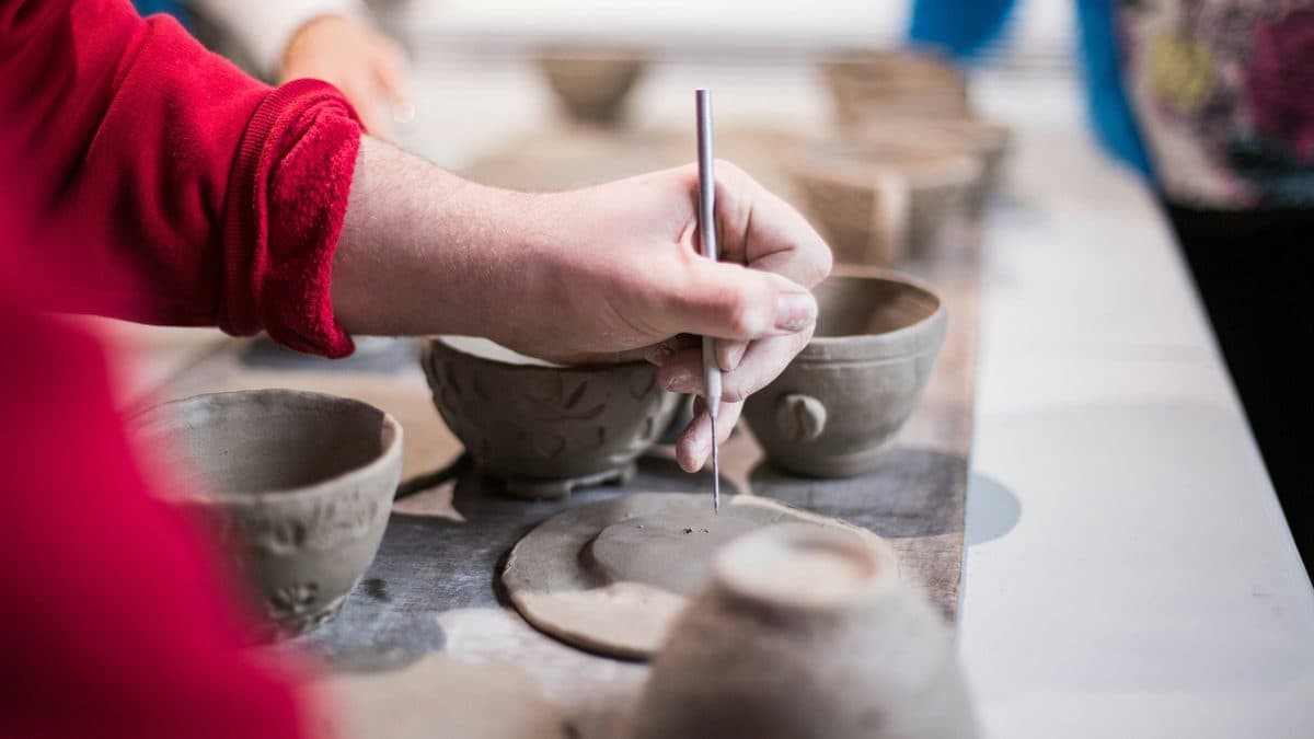 Hands carving a clay bowl, surrounded by hand-built pottery on a work table.